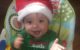 Baby boy wearing a red Santa hat and a green onesie that says "Santa's Little Helper." He has a huge smile and is clapping his hands together.