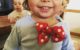 Two year old has a red hair bow with white polka dots on the front of his shirt, as if it was a bowtie. He's giggling about it.