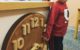 Almost three year old stands next to a big toy wooden clock with a big smile on his face.