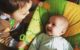 One year old looks at his baby sister in her froggy chair and they are both smiling big smiles at each other.