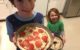 Seven year old big brother and 3 year old sister hold up their pizzas before putting them in the oven. They're smiling because they have pizza.