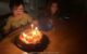 Middle kid smiles and looks at the candles on his jack-o-lantern birthday cake, while his smiling younger sister looks on